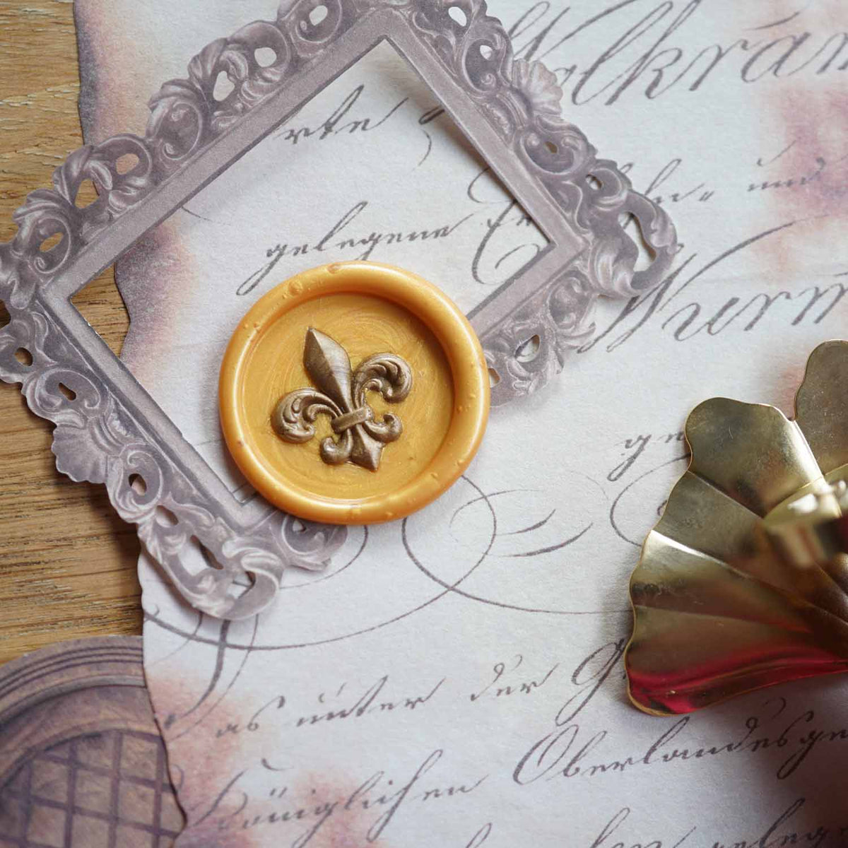 A vintage-style wax seal stamp set featuring a round golden wax seal with a fleur-de-lis motif, a translucent gray ornate frame die, and a gold metal stamp blade, arranged on aged script paper over a wooden surface.